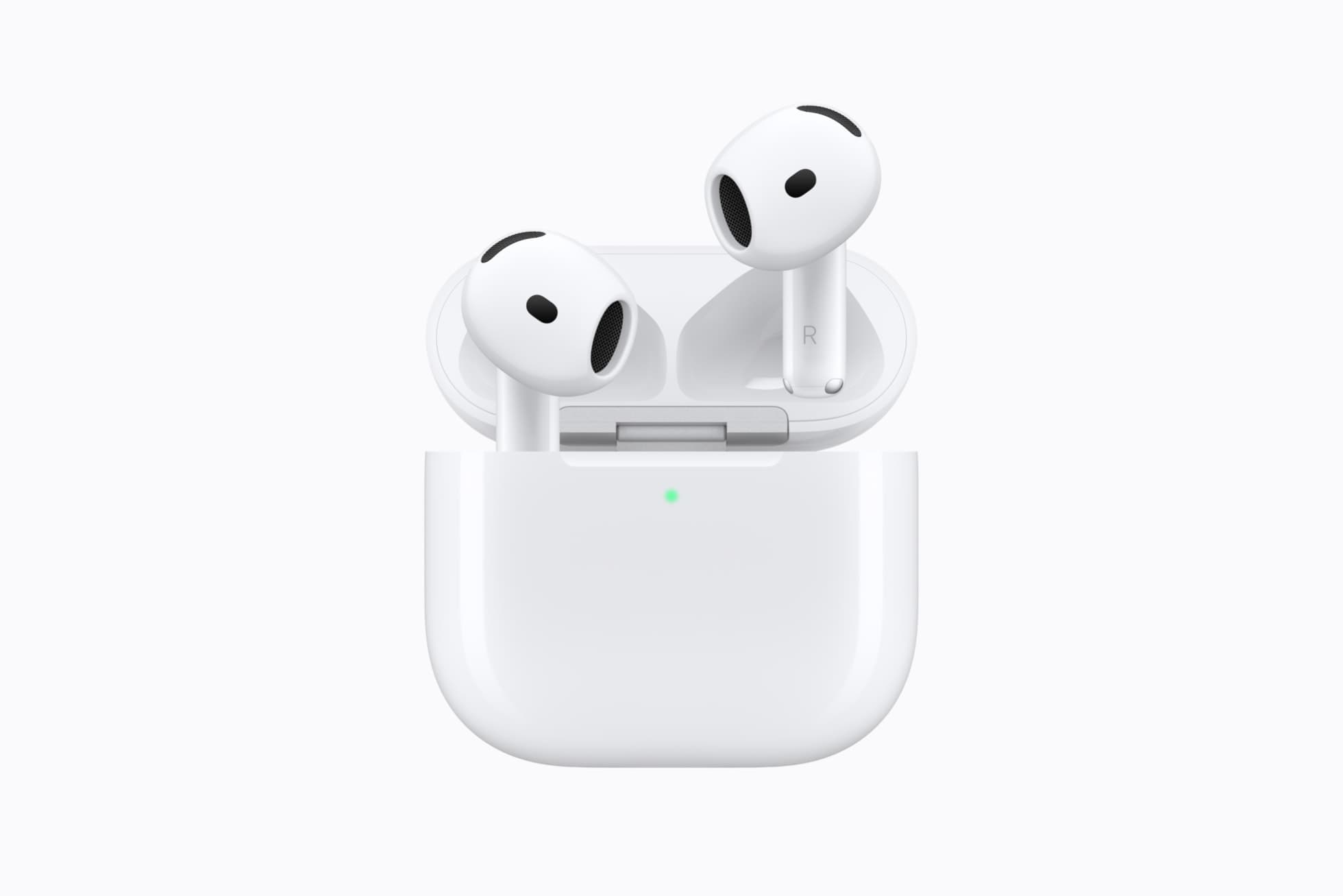 Two white AirPods 5 earbuds standing upright in their open white charging case, lid raised, with a small green status light glowing on the front of the case, against a pale gray background.