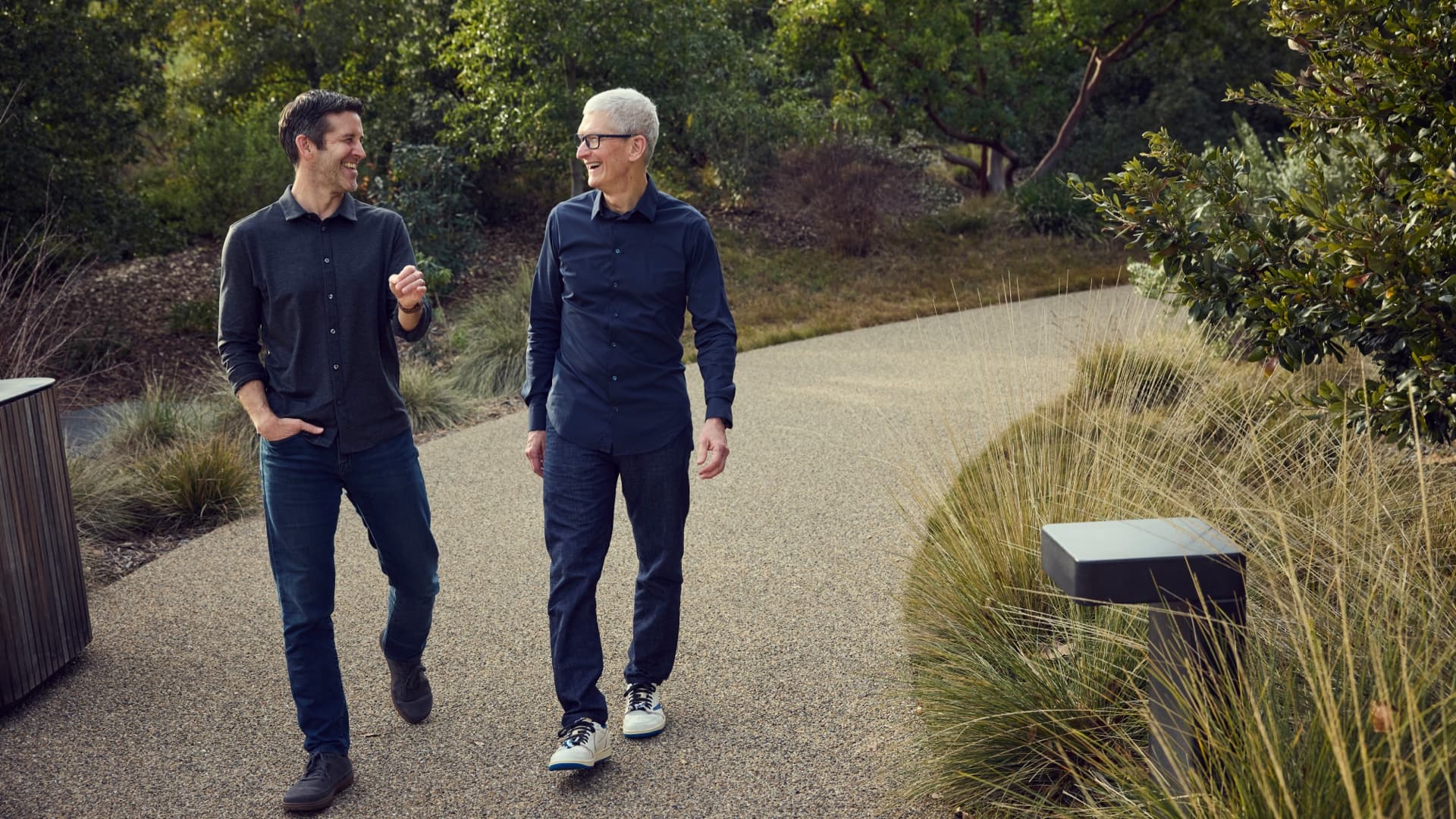 Two men in dark button-up shirts and jeans walk side by side down a curving gravel path between tall grasses and trees, turned toward each other and laughing, the one on the right in glasses and white sneakers; Apple captions the picture "Tim Cook and John Ternus at Apple Park."