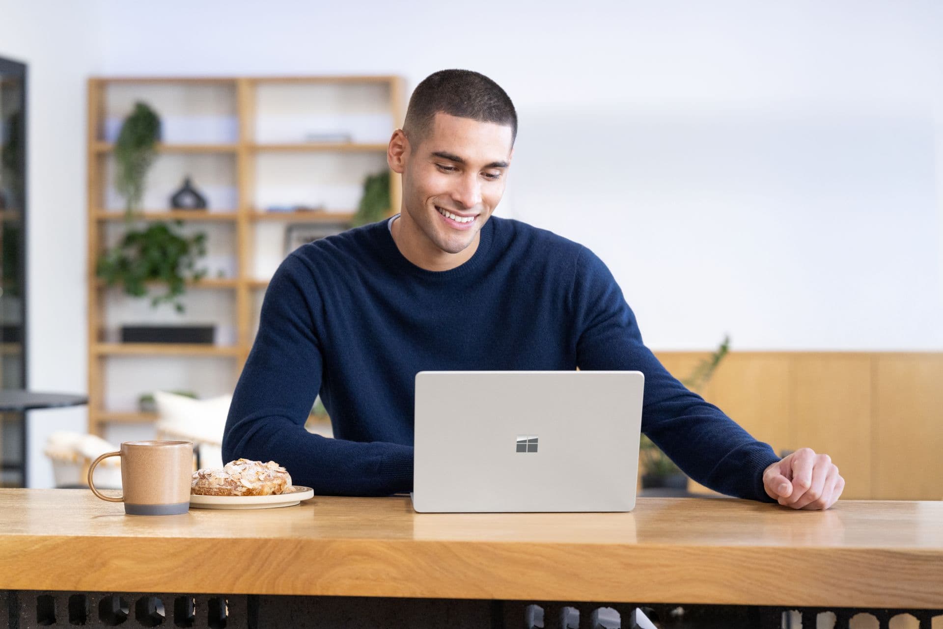 A smiling man in a navy sweater sits at a light wooden counter looking down at an open silver Surface laptop with the Microsoft logo on its lid; a tan mug and a pastry on a plate sit beside it, and wooden shelves with plants are out of focus behind him.