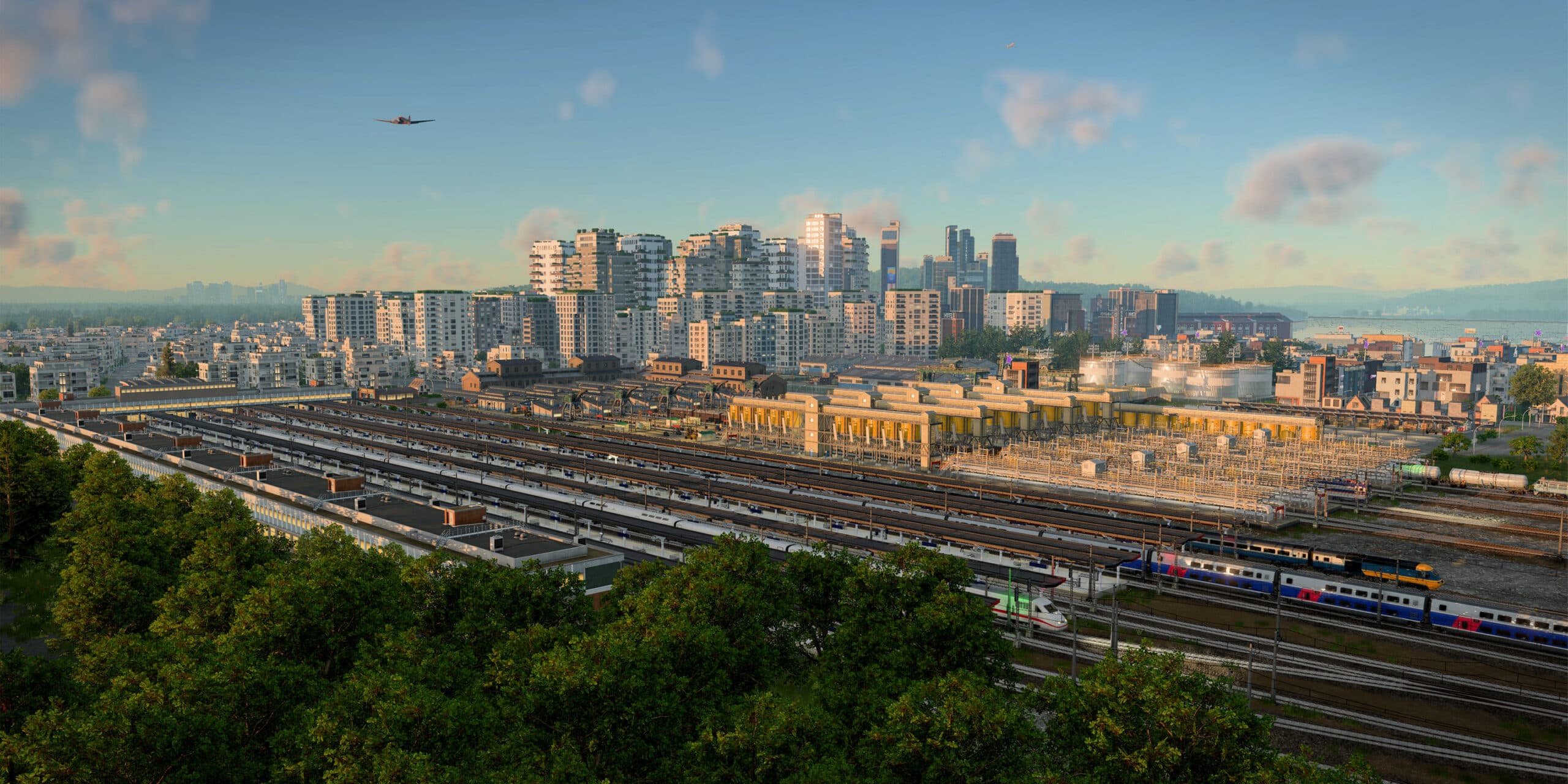 A wide in-game view over a busy railway station in warm evening light: trains stand under long platform canopies, with a white high-speed train and a blue, white and red passenger train headed by a blue and yellow locomotive at lower right, a yellow industrial plant, storage tanks and tank wagons behind the tracks, white apartment blocks and dark towers on the skyline, a propeller airliner in the sky at upper left and trees across the foreground.