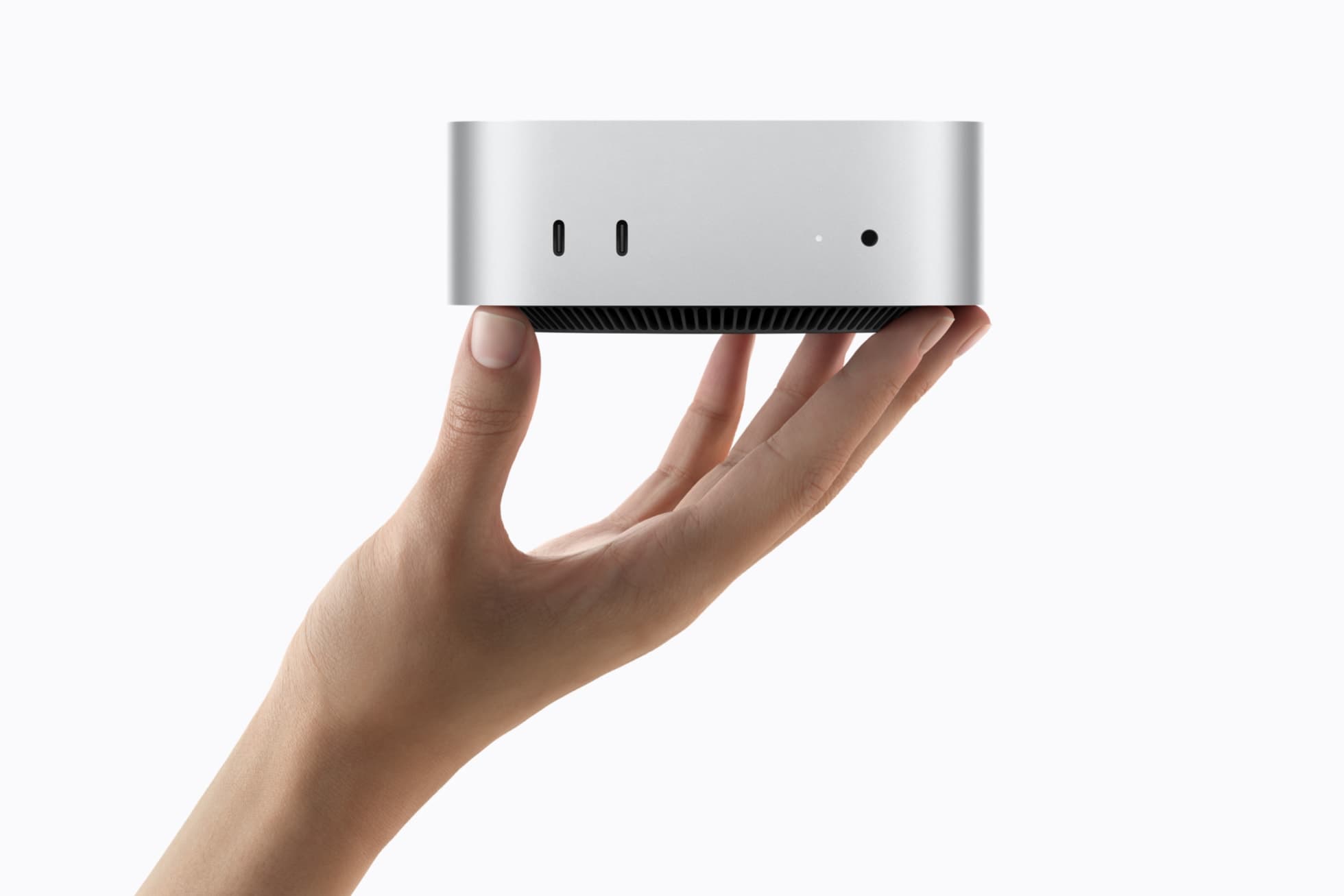 A hand holds a silver Mac mini flat on its fingertips against a white background, showing the front face with two USB-C ports, a small status light and a headphone jack above the black vented base.