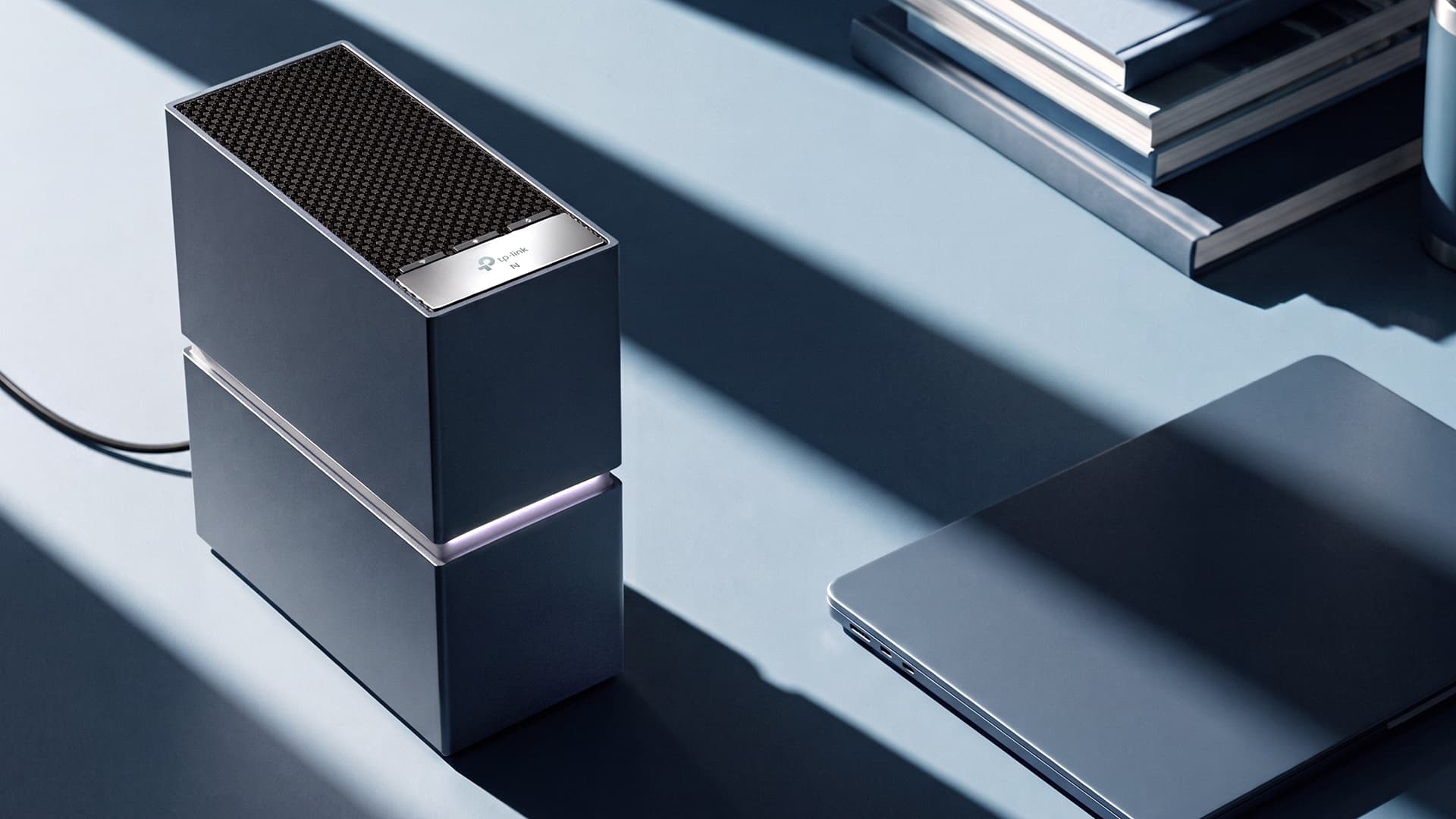 A dark slate-blue rectangular router stands upright on a pale blue surface crossed by bands of sunlight and shadow; a glowing seam divides it across the middle, its top has a patterned grille and a silver strip with the TP-Link logo, a cable trails off to the left, a closed laptop lies to its right and a stack of books sits behind.